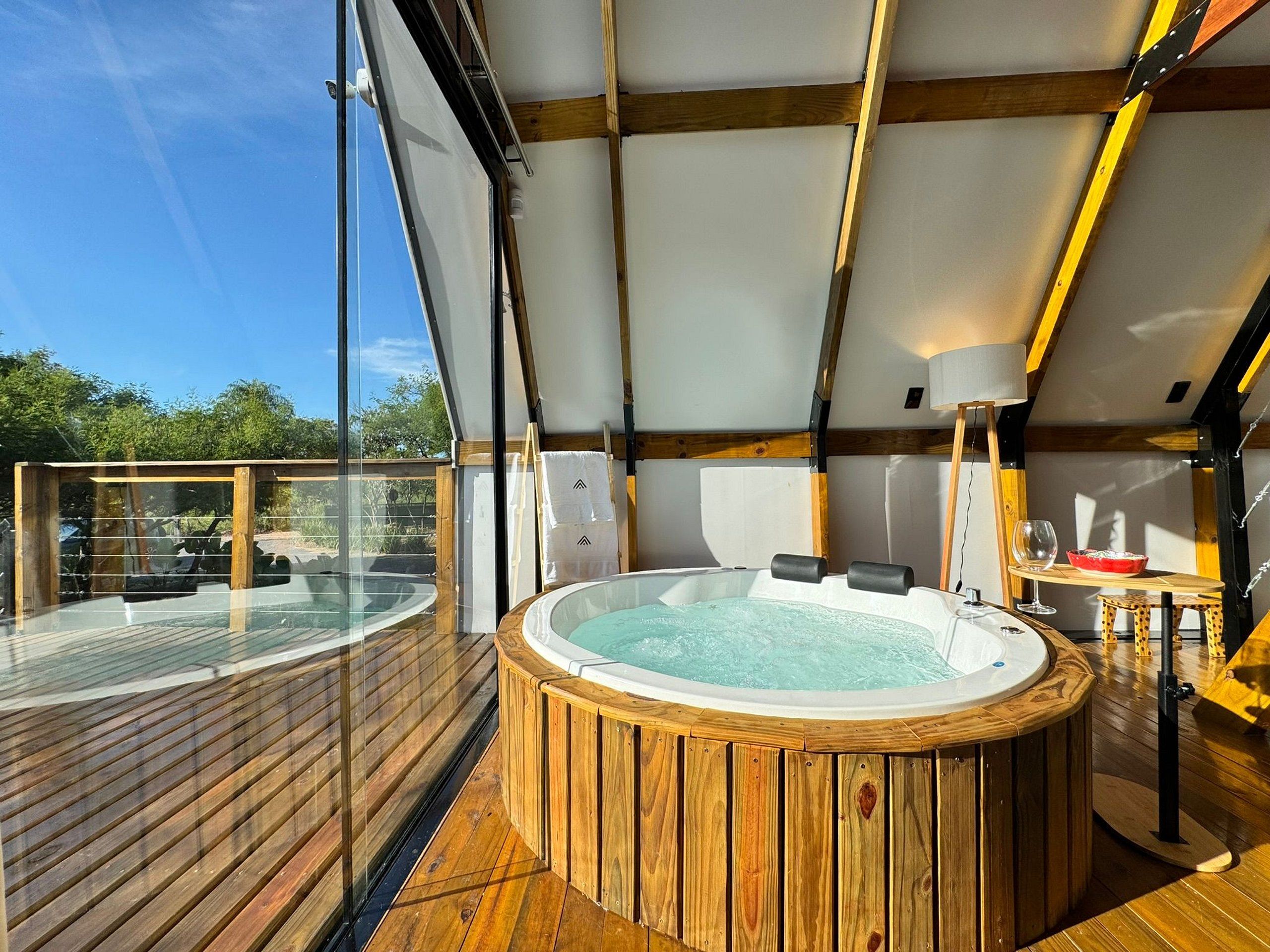 Un jacuzzi en una terraza de madera con barandilla de vidrio, vigas de madera y una vista despejada del cielo.