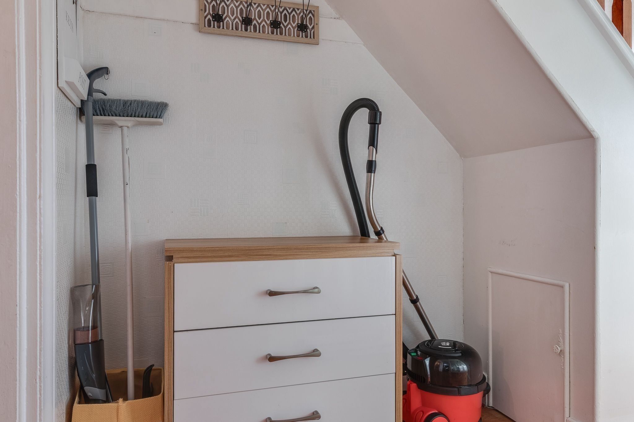 Et værelse med en hvid væg, et træskab med skuffer, en støvsuger og en kost, der læner sig op ad væggen.
