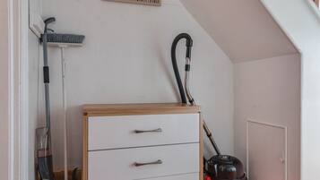Et værelse med en hvid væg, et træskab med skuffer, en støvsuger og en kost, der læner sig op ad væggen.