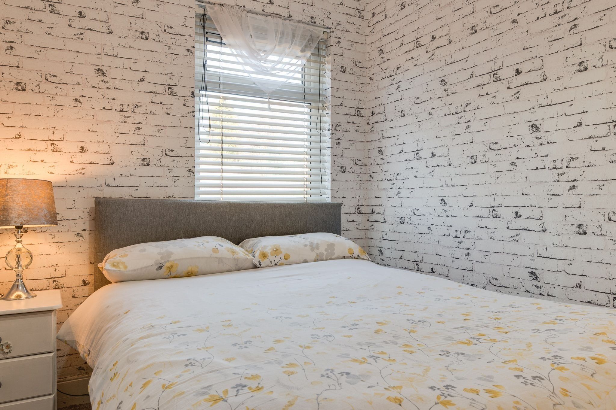 A bedroom with a brick-patterned wall, a bed with floral bedding, a bedside lamp, and a window with blinds.
