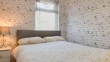 A bedroom with a brick-patterned wall, a bed with floral bedding, a bedside lamp, and a window with blinds.
