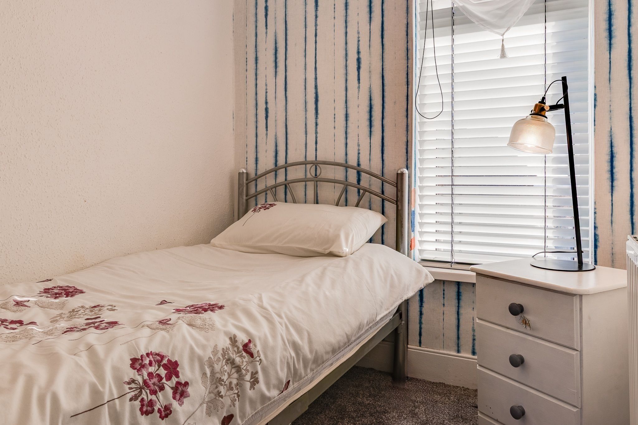 A single bed with a floral bedspread, a bedside table with drawers, a lamp, and a window with blinds.