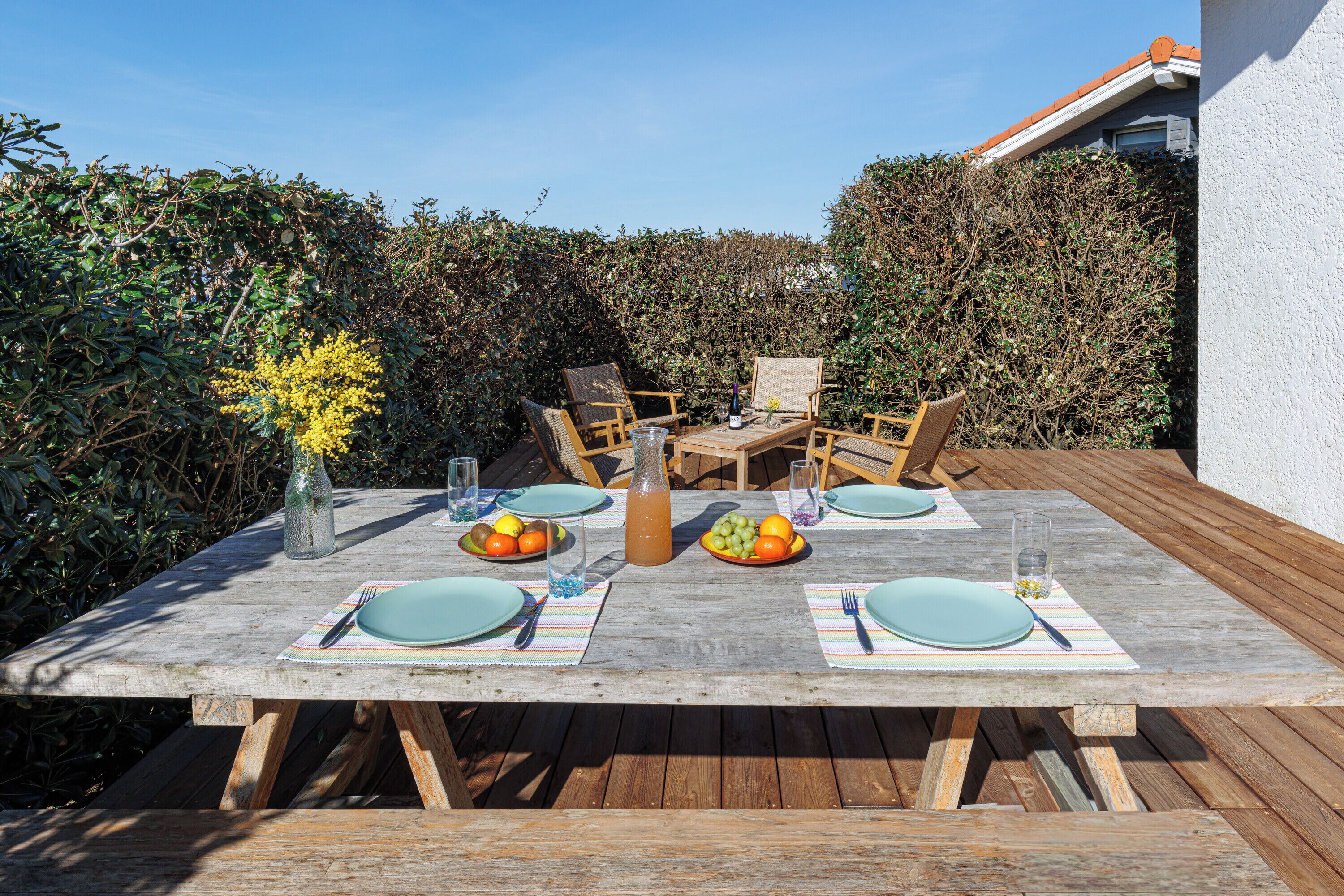 An outdoor dining area with a wooden table set for four, surrounded by chairs, with a hedge and a building in the background.