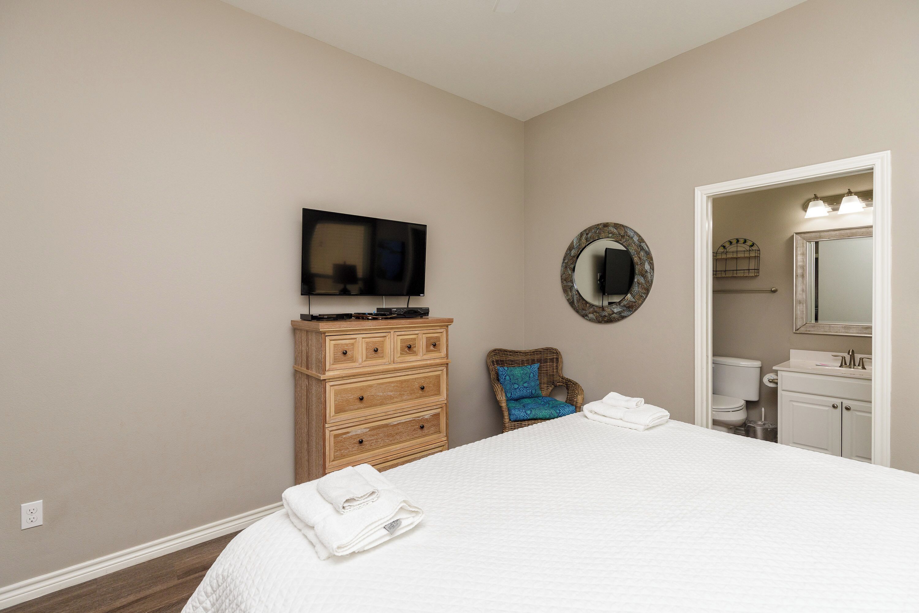 A bedroom with a bed, wooden dresser, television, wicker chair, and a bathroom visible through an open door.