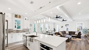 A modern kitchen with white cabinetry, a large island, and a dining area with a sofa and table. - Highland Parks, Resort-Style Pool! LSV/Golf Cart & 6 Bikes! (Santa Rosa Beach)