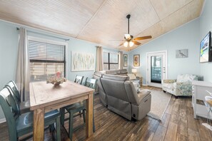 A spacious living area with a wooden ceiling, a ceiling fan, a grey sofa, a dining table with chairs, and a small white desk. - Lani Kai 230 – Gulf Coast Condo. Walk to Beach + Pools + Balcony Views! (Gulf Shores)