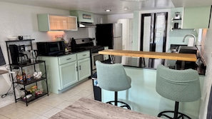 A compact kitchen with a built-in stove, a refrigerator, and a coffee machine. There are two bar stools and a small bar counter. - Home Away From Home-Fenced Yard-Firepit-Dogs OK-Close to Beach/Cruise Terminals (Galveston)