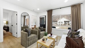 Un salon moderne avec un canapé, des fauteuils et une table basse. Il y a une grande fenêtre avec des rideaux, une porte qui donne sur une autre pièce et une vue sur une maison à l’extérieur.