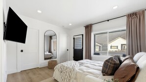 Une chambre à coucher comprenant un lit, un téléviseur à écran plat, un miroir, une fenêtre avec des rideaux et une vue sur une maison à l’extérieur.
