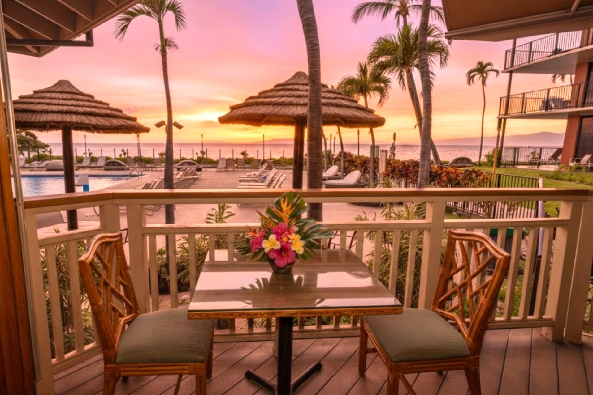 A balcony with two wicker chairs and a table, overlooking a pool area with thatched umbrellas and lounge chairs, set against a sunset view.