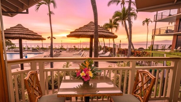 A balcony with two wicker chairs and a table, overlooking a pool area with thatched umbrellas and lounge chairs, set against a sunset view. - Kaanapali Shores | Ocean View Studio Sleeps 4 | Car Incl. w/ 6+ Nights | KS-736 by KBM (Lahaina)