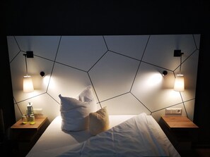 A modern hotel room with a geometric-patterned headboard, two bedside tables with lamps, and a neatly made bed with pillows. - Conference Partner Ullrich (Elfershausen)