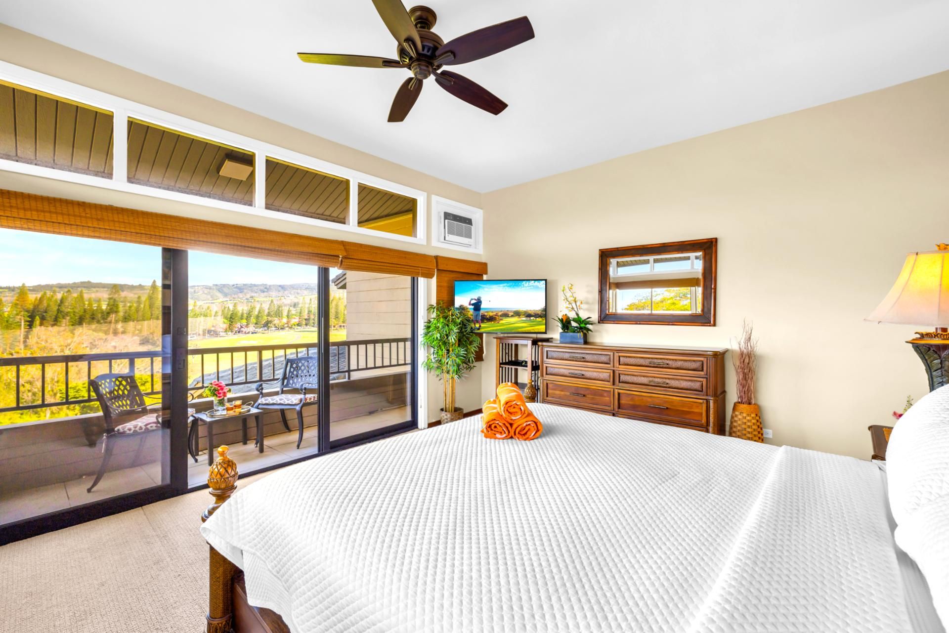 A bedroom with a large bed, a ceiling fan, a TV, a dresser, and a balcony with a view.