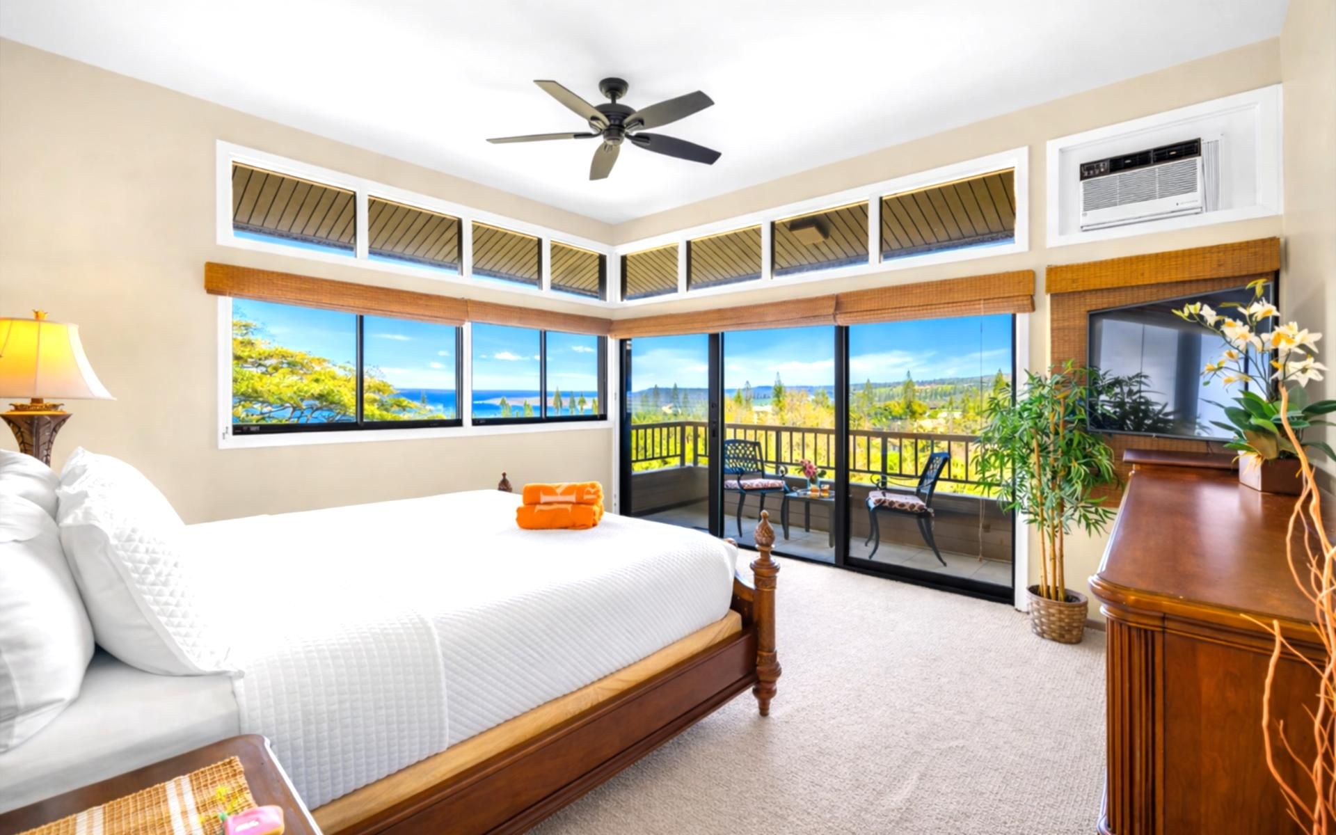 A bedroom with a large bed, a ceiling fan, a view of the outdoors, and a balcony with chairs.