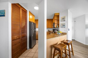 A kitchen with wooden cabinets, a refrigerator, and a bar area with stools. - Kaanapali Shores | Ocean View 3 BR, Sleeps 10 | Car Incl. w/ 6+ Nights | KS ML-3659 by KBM (Lahaina)