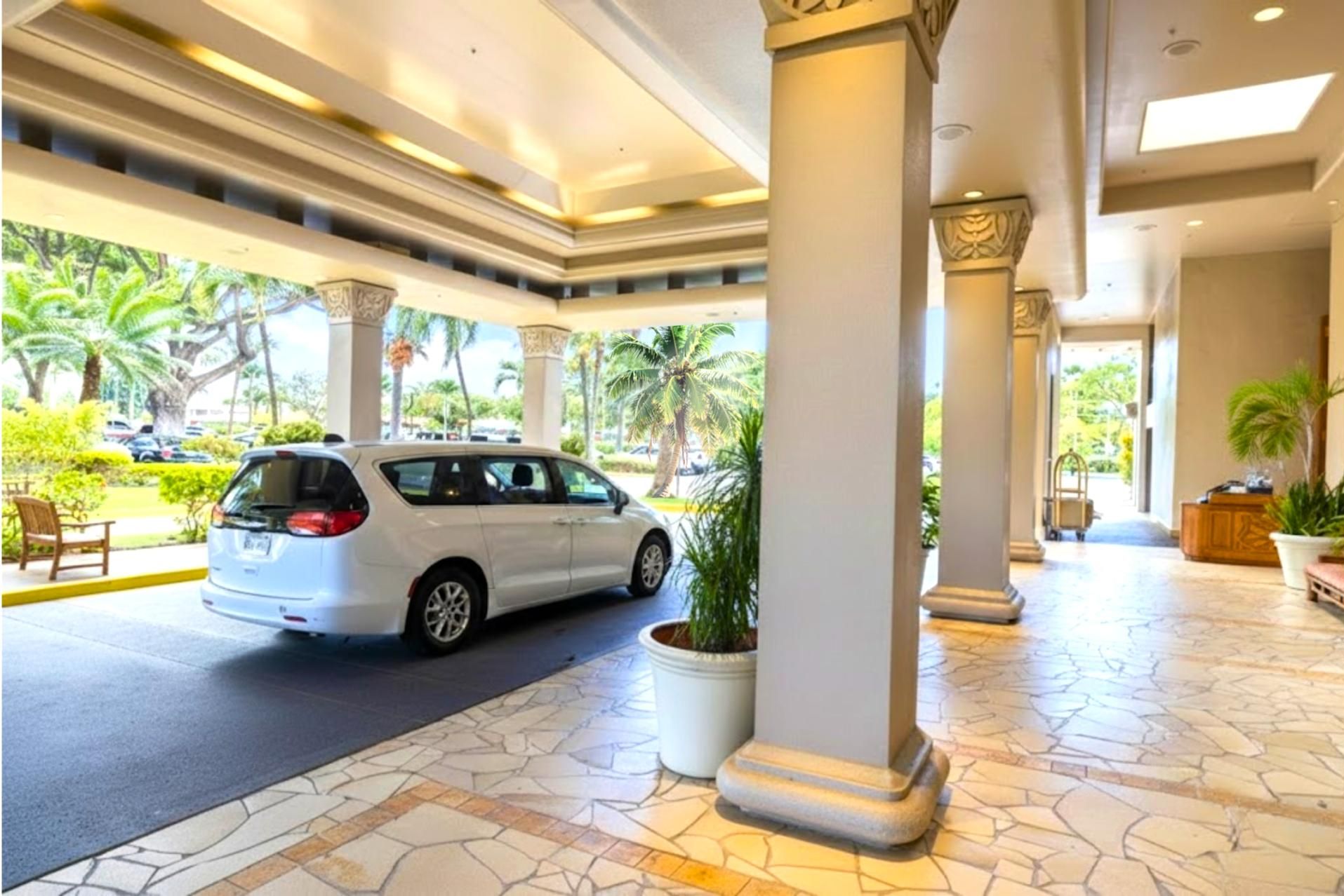 A white car parked in a covered area with columns and a tiled floor, surrounded by greenery and palm trees.