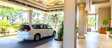 A white car parked in a covered area with columns and a tiled floor, surrounded by greenery and palm trees.