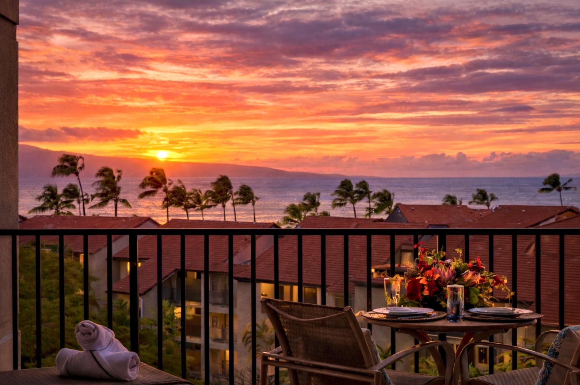 Un balcon avec une table dressée pour deux personnes, offrant une vue sur un coucher de soleil sur l’océan, avec des palmiers en arrière-plan.
