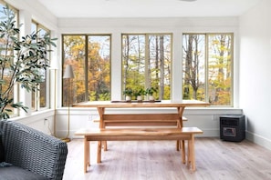A room with a wooden bench, a table, and three large windows offering a view of trees.