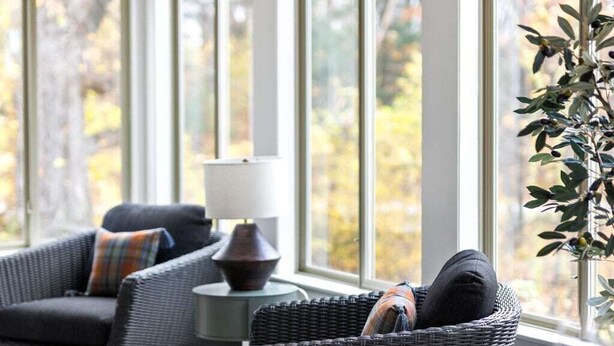 Ein gemütliches Zimmer mit Korbmöbeln, einem Tisch mit Tellern und einem Glas sowie großen Fenstern mit Blick auf Bäume.