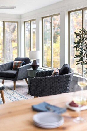 A cozy room with wicker furniture, a table with plates and a glass, and large windows with a view of trees. - Devils Dive · Devils Dive Estate (Traverse City)