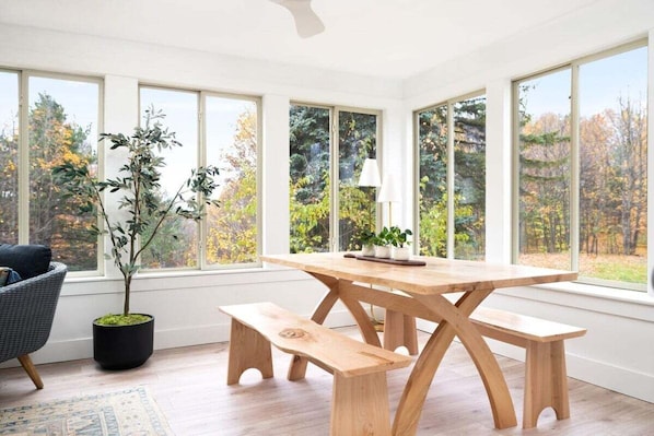 A dining area with a wooden table and benches, large windows with a view of trees, and a potted plant.