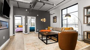 Ein modernes Wohnzimmer mit einer Couch, einem Couchtisch und einem Kamin. Zwei Türen führen in andere Räume, und ein großes Fenster sorgt für natürliches Licht.