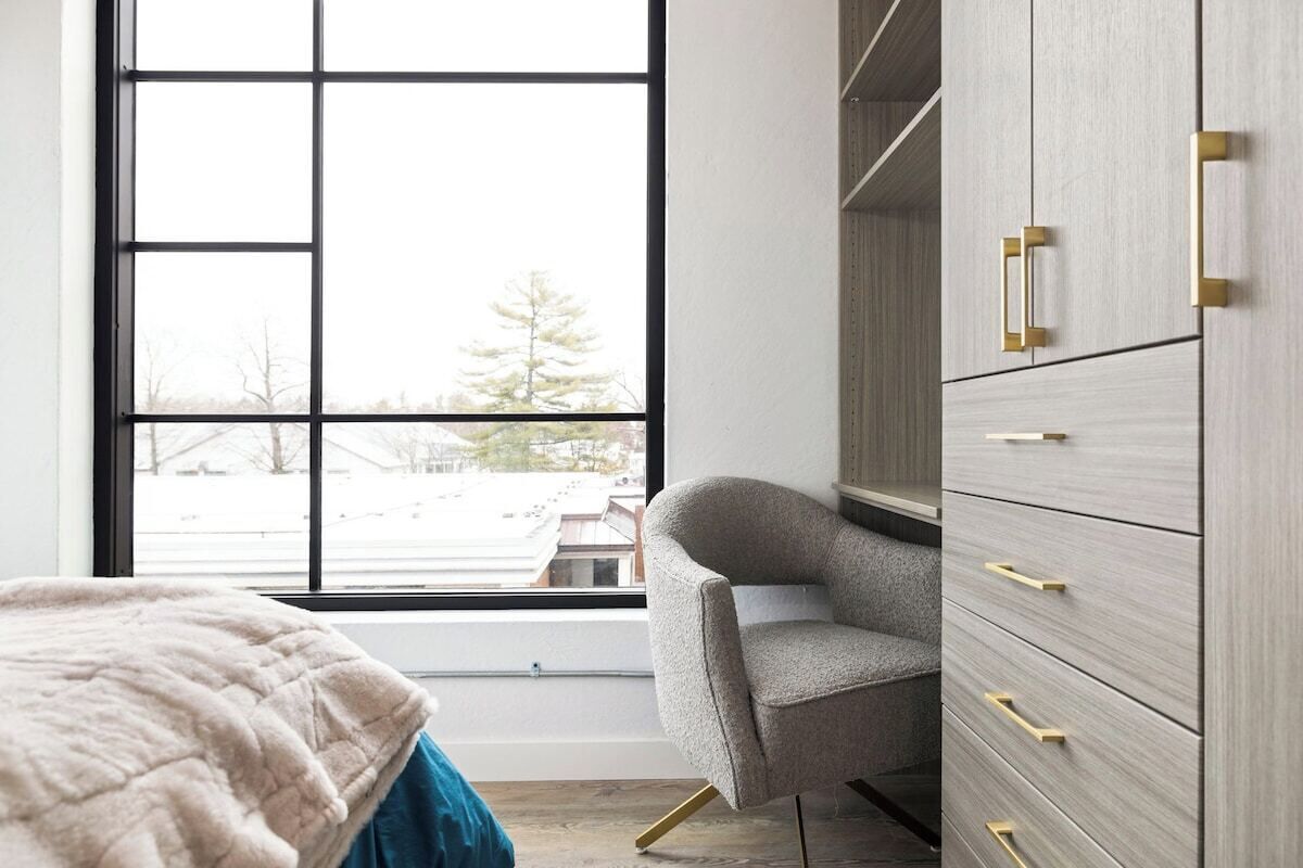 A bedroom with a large window, a bed with a blue blanket, a grey armchair, and a grey wardrobe with gold handles.