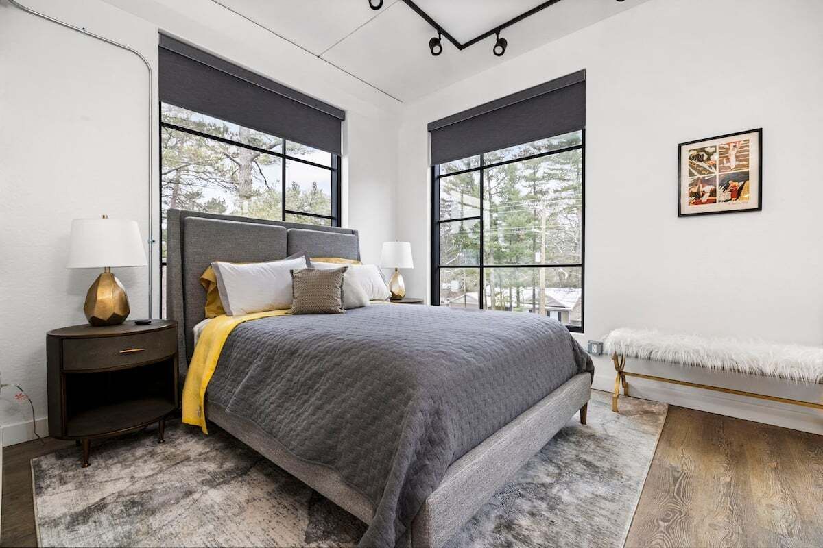 Une chambre moderne avec un grand lit, deux tables de chevet, un banc et de grandes fenêtres donnant sur la forêt.