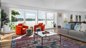 A spacious living room with large windows, a glass coffee table, and a grey sofa with patterned cushions. - Beach House - Lakefront with a Hot Tub! (Traverse City)