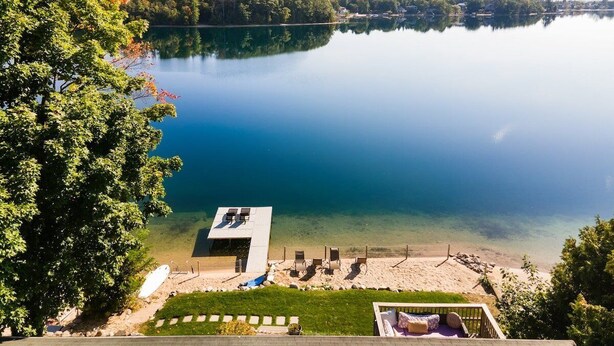 A lakeside view with a dock, a small building with a roof, and a grassy area with chairs. - Beach House - Lakefront with a Hot Tub! (Traverse City)
