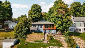 A modern two-story house with a covered patio, a stone retaining wall, and a well-maintained lawn. - Beach House - Lakefront with a Hot Tub! (Traverse City)