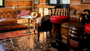 A rustic cabin interior with a stone-tiled floor, wooden walls, a leather couch, a red and black chair, and a large beer barrel.