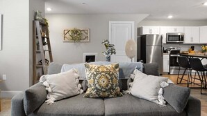 A modern living room with a grey sofa, decorative pillows, a wooden ladder, and a kitchen area with white cabinets and stainless steel appliances. - TC Parkside H: Charming 2BR Retreat (Traverse City)