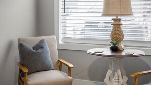 A room with a beige armchair, a round table with a lamp, and a window with blinds. - TC Parkside H: Charming 2BR Retreat (Traverse City)