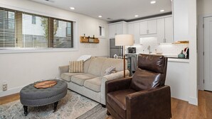 Ein modernes Wohnzimmer mit Sofa, Hocker und Ledersessel, eine Küche mit weißen Schränken und ein großes Fenster mit Jalousien.