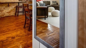 Een modern interieur met openliggende bakstenen muren, houten vloeren, en een grote spiegel die een zithoek met een bank, voetenbankje en een eettafel weerspiegelt.