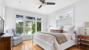 Ein Schlafzimmer mit einem weißen Bett, einem Schreibtisch und einem Stuhl. Es gibt zwei Fenster und einen Deckenventilator.