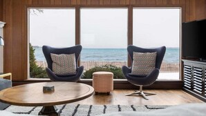 Een kamer met een groot raam met uitzicht op zee, twee fauteuils met gekleurde kussens, een klein voetenbankje, een ronde houten tafel en een televisie op een kast.