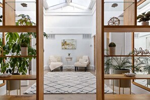 A modern living room with two armchairs, a coffee table, and a geometric rug. There are potted plants and a painting on the walls.