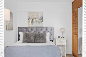 A bedroom with a tufted headboard, a grey and white abstract painting, a bedside lamp, and a wooden door.
