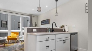 Eine moderne Küche mit weißen Schränken, einer grauen Arbeitsplatte und einem eingebauten Geschirrspüler. Angrenzend ein Wohnbereich mit einem gelben Stuhl und einem kleinen Tisch.