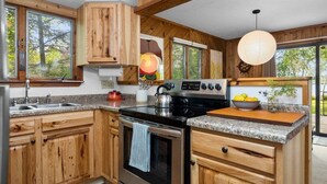 A modern kitchen with wooden cabinets, a stainless steel stove, granite countertops, and large windows offering a view of trees. - Camp Birch: Waterfront Home on Lake Leelanau (Lake Leelanau)