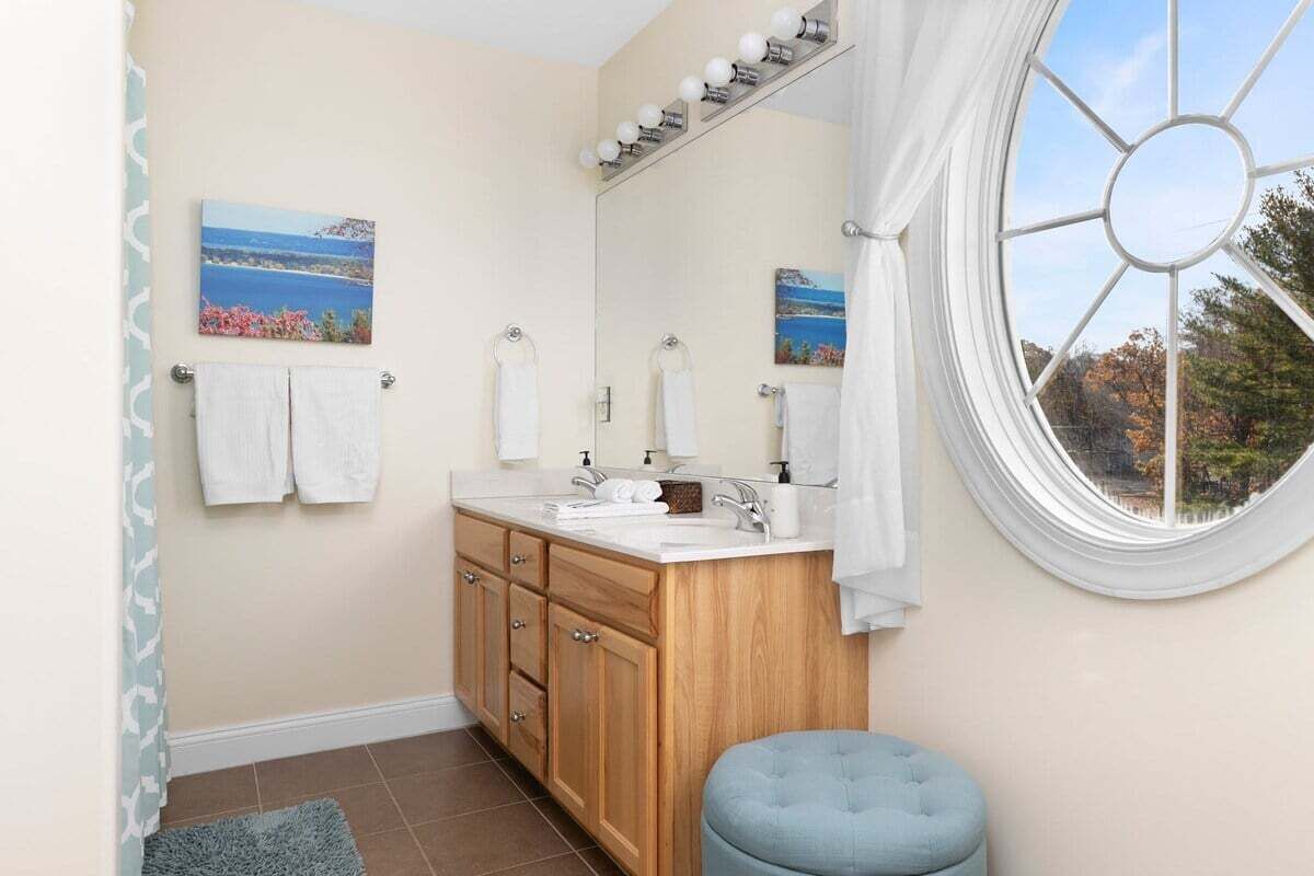 Een badkamer met een houten wastafelmeubel, een grote spiegel en een raam met uitzicht op bomen.