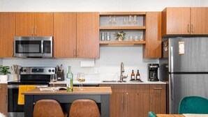 Una cocina moderna con armarios de madera, un refrigerador de acero inoxidable, un microondas y una cafetera. Hay dos taburetes de bar y una pequeña isla con una copa de vino y una botella.