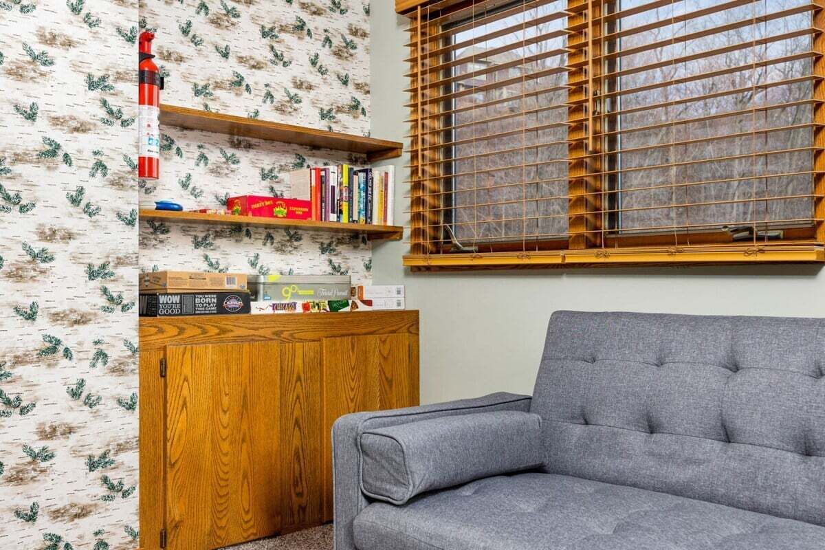 A room with a grey sofa, wooden shelves with books and toys, a wooden cabinet, and a window with wooden blinds.