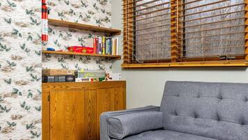 A room with a grey sofa, wooden shelves with books and toys, a wooden cabinet, and a window with wooden blinds.