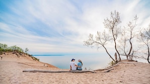 Une plage de sable où un couple est assis sur une bûche, surplombant une étendue d’eau calme et quelques arbres clairsemés.
