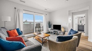 Una sala de estar moderna con un sofá, sillones, una mesa de centro y un televisor. Hay una puerta corrediza de vidrio que da a un balcón.