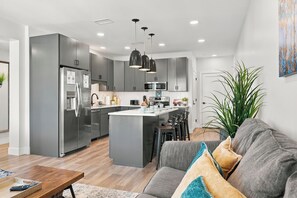 A modern kitchen with grey cabinets, a central island, and a dining area with a grey sofa and a coffee table. - Cityscape Condo: New in Downtown Traverse City! (Traverse City)